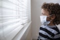 Young boy looking out the window wearing a protective facemark while seeking protection from COVID-19, or the novel coronavirus