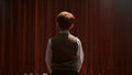 A young boy in a school uniform stands on a stage his back to the camera as he recites a monologue with passion and