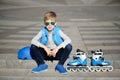 Young boy sitting near grey wall with inline roller skates and all protection at outdoor skate park Royalty Free Stock Photo