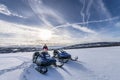 Young boy stands behind two blue snomobiles in winter snowy mountains, cold virgin snow, bright sun with halo around, Norway