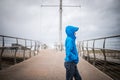 Young boy stood on bridge looking out to sea away from camera hood up in blue coat rain storm weather and wind blowing outdoors Royalty Free Stock Photo