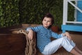 Young boy in the striped shirt is sitting next to a wooden boat and dreamily looks into the distance. Studio shot Royalty Free Stock Photo