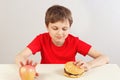 Young boy at the table chooses between hamburger and fruit on white background