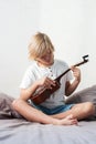 Young boy tuning ukulele at home. Blond haired boy sitting on couch playing acoustic guitar
