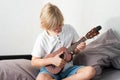 Young boy tuning ukulele at home. Blond haired boy sitting on couch playing acoustic guitar Royalty Free Stock Photo