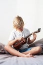 Young boy tuning ukulele at home. Blond haired boy sitting on couch playing acoustic guitar Royalty Free Stock Photo