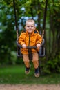 Young boy wearing orange raincoat smiles in swings in the park, cheerful childhood