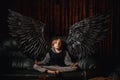 A young boy, 9-10 years old, sits on a couch in a dark room, meditating with his black angel wings spread wide. The boy looks Royalty Free Stock Photo