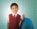 Young boy 7 or 8 years old in vest and necktie uniform holding bag smiling cheerful in front of green chalkboard excited about Royalty Free Stock Photo