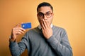 Young brazilian businessman holding credit card money over isolated yellow background cover mouth with hand shocked with shame for Royalty Free Stock Photo