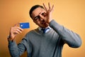 Young brazilian businessman holding credit card money over isolated yellow background with happy face smiling doing ok sign with Royalty Free Stock Photo