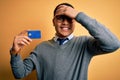 Young brazilian businessman holding credit card money over isolated yellow background stressed with hand on head, shocked with Royalty Free Stock Photo