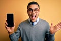 Young brazilian businessman wearing glasses holding smartphone showing screen very happy and excited, winner expression Royalty Free Stock Photo