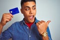 Young brazilian customer man holding credit card standing over isolated white background pointing and showing with thumb up to the Royalty Free Stock Photo