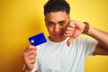 Young brazilian customer man holding credit card standing over isolated yellow background with angry face, negative sign showing Royalty Free Stock Photo