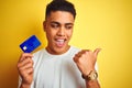 Young brazilian customer man holding credit card standing over isolated yellow background pointing and showing with thumb up to Royalty Free Stock Photo