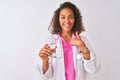 Young brazilian doctor woman holding glass of water standing over isolated white background with surprise face pointing finger to Royalty Free Stock Photo