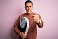 Young brazilian man doing diet to lose weigth holding scale over isolated pink background annoyed and frustrated shouting with