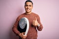 Young brazilian man doing diet to lose weigth holding scale over isolated pink background happy with big smile doing ok sign,