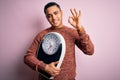 Young brazilian man doing diet to lose weigth holding scale over isolated pink background doing ok sign with fingers, excellent