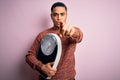 Young brazilian man doing diet to lose weigth holding scale over isolated pink background pointing with finger to the camera and