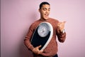 Young brazilian man doing diet to lose weigth holding scale over isolated pink background pointing and showing with thumb up to