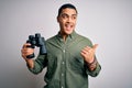 Young brazilian man looking through binoculars over isolated white background pointing and showing with thumb up to the side with Royalty Free Stock Photo