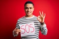 Young brazilian man with problem holding banner with sos message over red background doing ok sign with fingers, excellent symbol