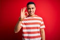 Young brazilian man wearing casual striped t-shirt standing over isolated red background smiling positive doing ok sign with hand Royalty Free Stock Photo