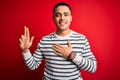 Young brazilian man wearing casual striped t-shirt standing over isolated red background smiling swearing with hand on chest and Royalty Free Stock Photo