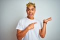 Young brazilian man wearing king crown standing over isolated white background amazed and smiling to the camera while presenting