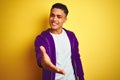 Young brazilian man wearing purple sweatshirt standing over isolated yellow background smiling friendly offering handshake as