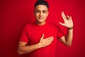 Young brazilian man wearing t-shirt standing over isolated red background smiling swearing with hand on chest and fingers up, Royalty Free Stock Photo
