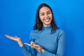 Young brazilian woman standing over blue isolated background amazed and smiling to the camera while presenting with hand and