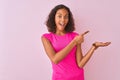 Young brazilian woman wearing t-shirt standing over isolated pink background amazed and smiling to the camera while presenting