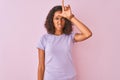 Young brazilian woman wearing t-shirt standing over isolated pink background making fun of people with fingers on forehead doing Royalty Free Stock Photo