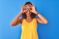 Young brazilian woman wearing yellow t-shirt standing over isolated blue background doing ok gesture like binoculars sticking Royalty Free Stock Photo