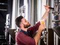 Young brewer wearing a leather apron is testing beer at a modern brewery