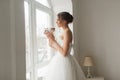 Young bride in a beautiful dress holding a bouquet of flowers posing near window in bright white studio. Wedding concept