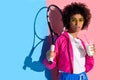 Young bright african american girl holding tennis racket and plastic cup with drink on pink and blue Royalty Free Stock Photo