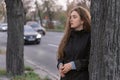 Young brooding woman stands by tree near highway on cloudy autumn day. Teen girl in coat on road background Royalty Free Stock Photo