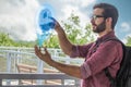 A young brown hair bearded man wearing glasses clicks a hologram projection of two palm trees shooting out of his phone outdoors