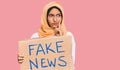 Young brunette arab woman holding fake news banner serious face thinking about question with hand on chin, thoughtful about