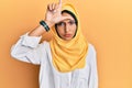 Young brunette arab woman wearing traditional islamic hijab scarf making fun of people with fingers on forehead doing loser Royalty Free Stock Photo
