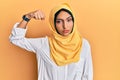 Young brunette arab woman wearing traditional islamic hijab scarf strong person showing arm muscle, confident and proud of power