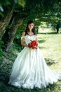 Young brunette bride with beautiful smile is holding the red wedding bouquet. Park location. Vertical full-length shot.