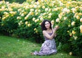 Young brunette Caucasian woman with curly hair sitting on green grass near yellow roses Bush in a garden, looking straight to the Royalty Free Stock Photo