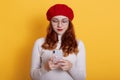 Young brunette Caucasian woman holds modern mobile phone, sends messages, reads funny notification, wears red beret, white shirt Royalty Free Stock Photo