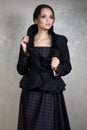 Young brunette with collected hair in a dark plaid dress and jacket with high collar standing on gray background Royalty Free Stock Photo