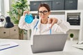 Young brunette doctor woman holding alarm clock with angry face, negative sign showing dislike with thumbs down, rejection concept Royalty Free Stock Photo